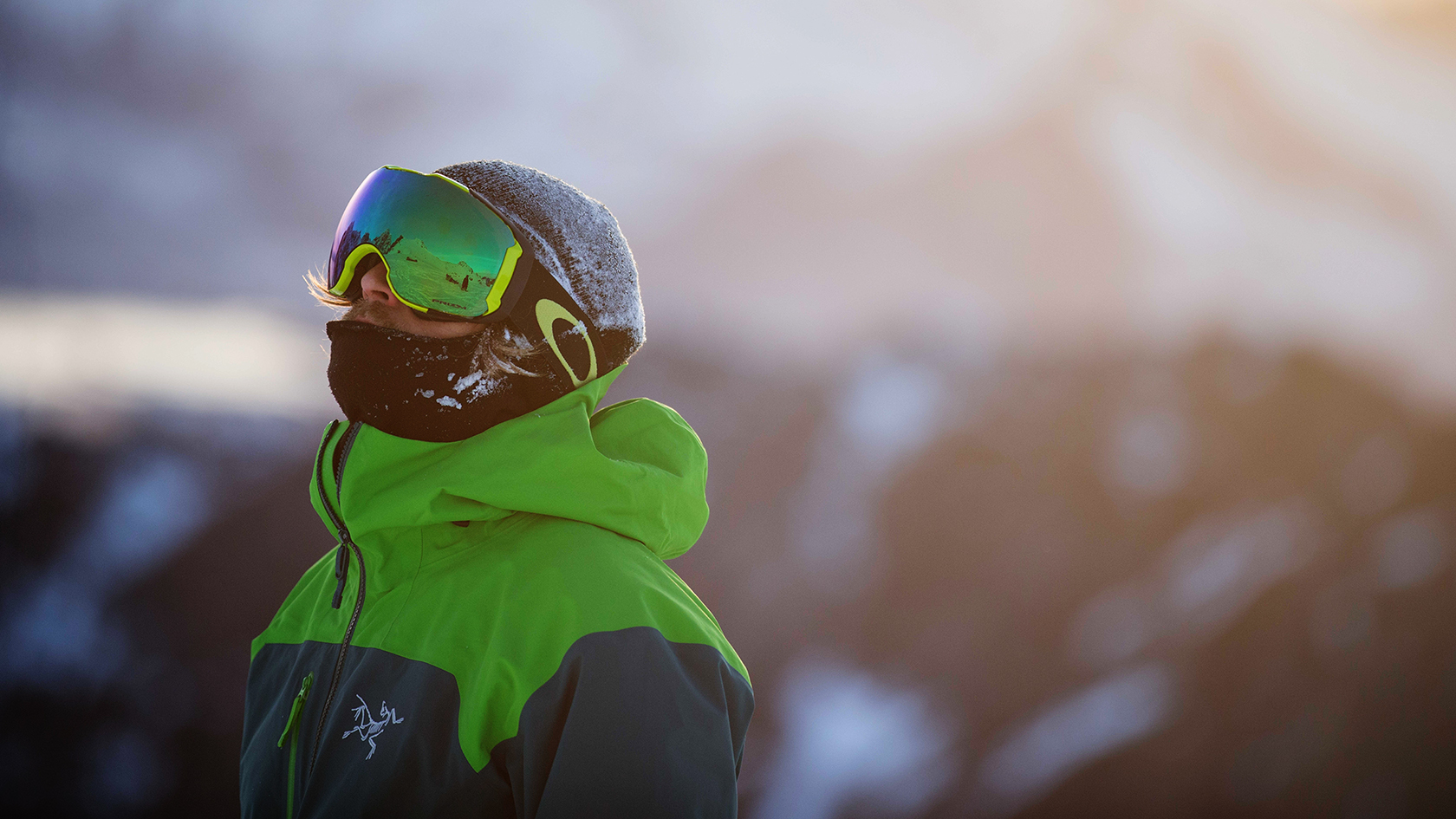 A man in a bright orange ski jacket holding various Oakley Prizm goggles is talking to the camera