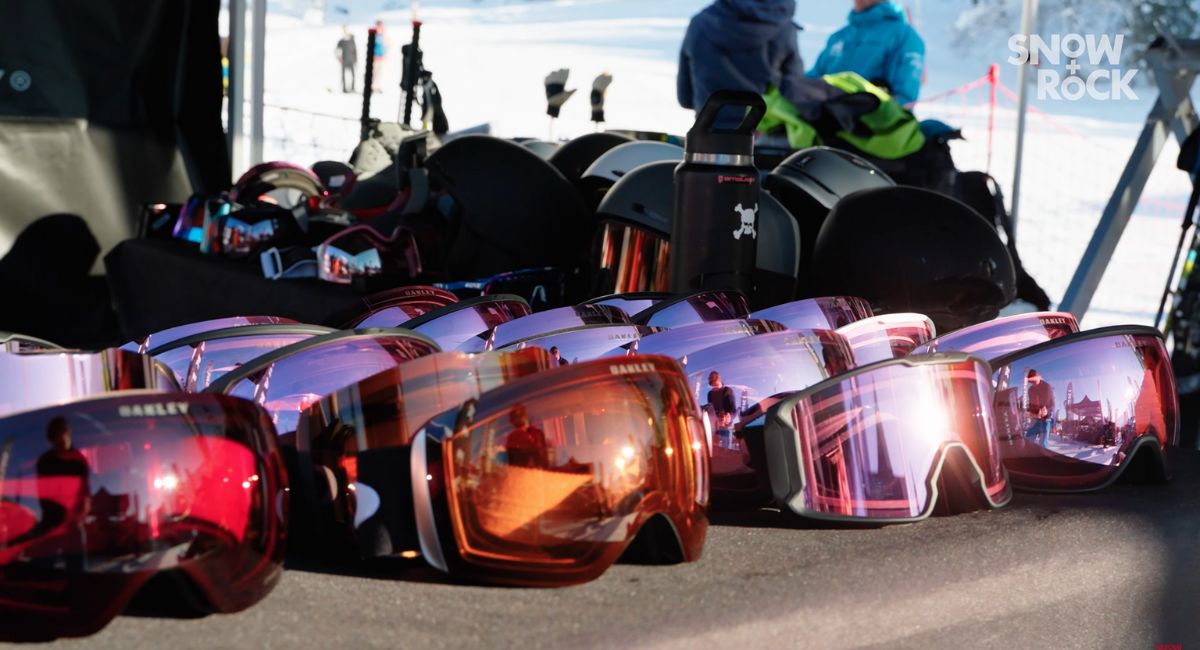 A man in a bright orange ski jacket holding various Oakley Prizm goggles is talking to the camera