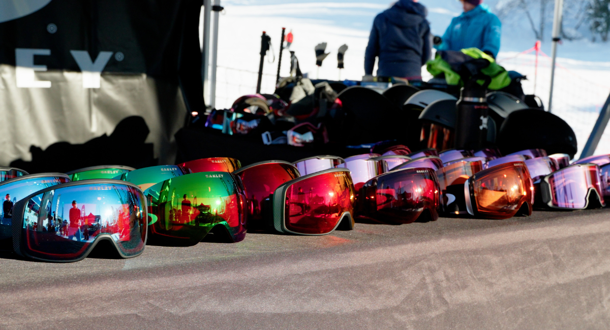 A man in a bright orange ski jacket holding various Oakley Prizm goggles is talking to the camera