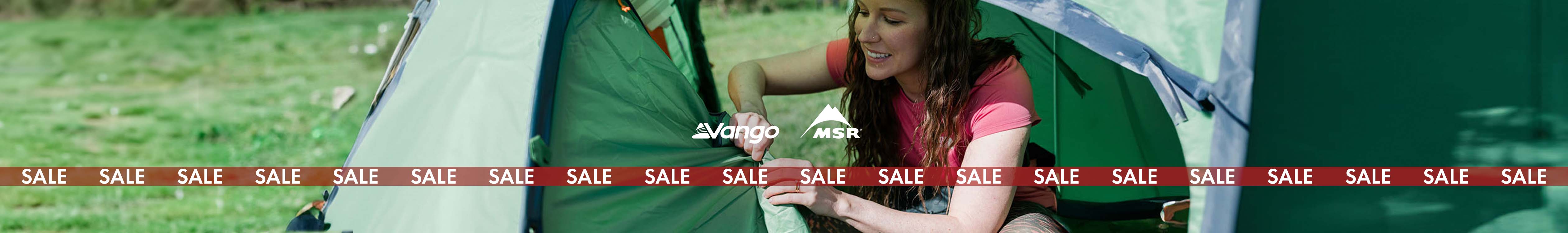 Camping sale. A woman is setting us a green tent, wearing a pink t-shirt 