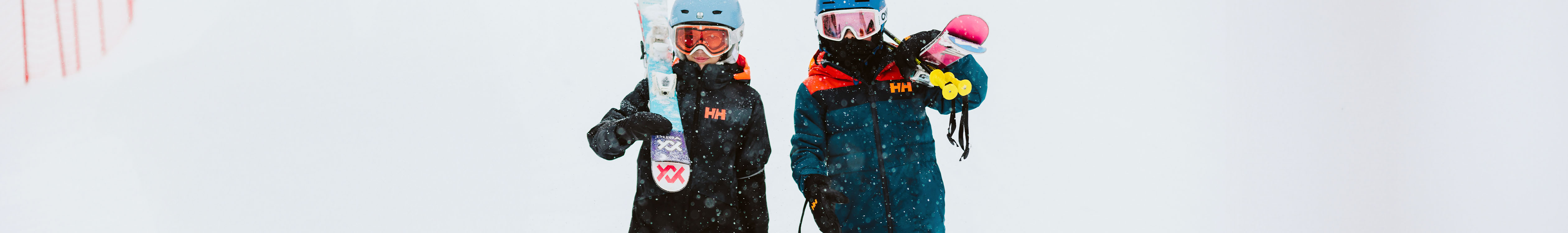 Shop Kids jackets. Two kids in Helly Hansen jackets standing outside in a snow holding skies