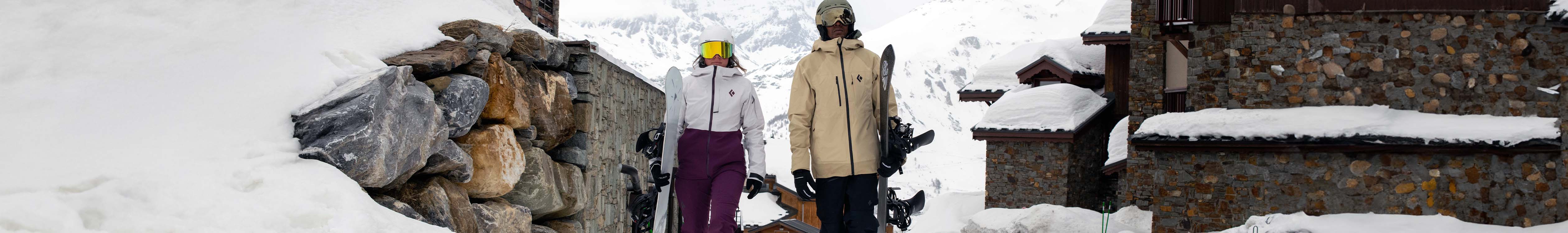 Shop Snowboard clothing. Two people holding their snowboards in snowsports clothing in front of a chalet