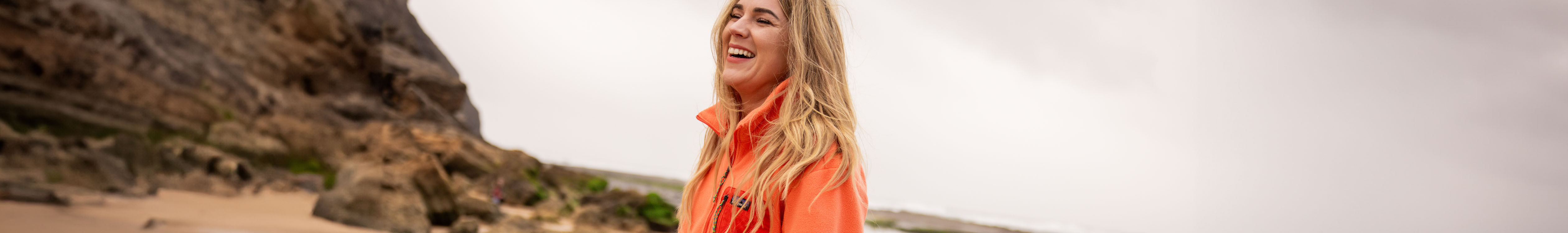 Fleeces summer styles. A woman is on a beach, wearing an orange fleece and smiling