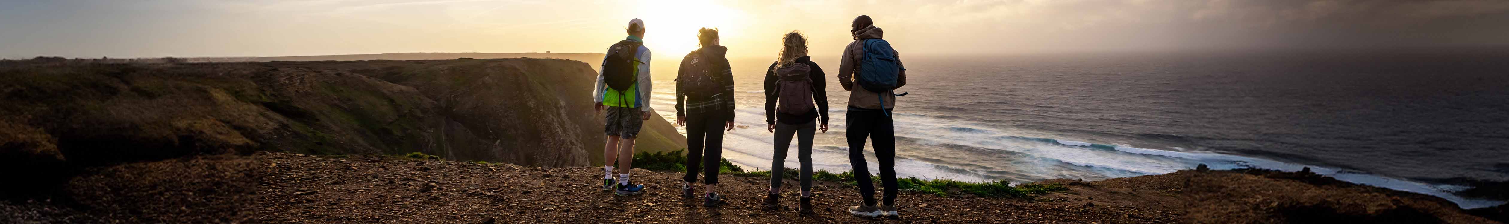 Offers.  4 People are looking at the going down sun and the sea, wearing hiking gear.