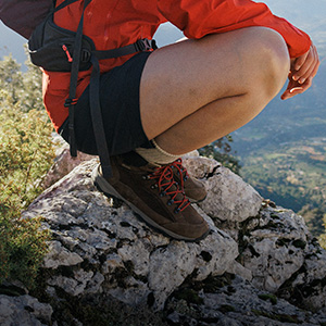 Shop walking boots + shoes. A man in shorts and walking boots squatting on a rock