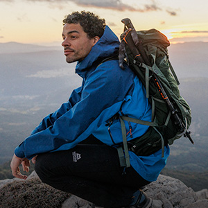 Shop jackets. A man in a blue jacket carrying a backpack is resting during a mountain hike