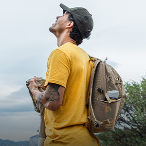 Shop summer accessories. A man wearing yellow T-shirt and a khaki cap, and carrying a khaki backpack is standing on a mountain