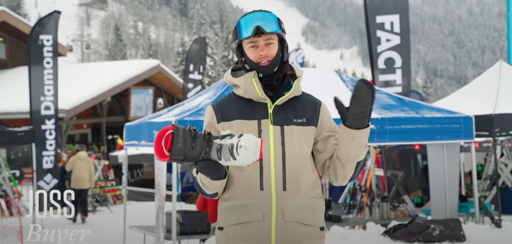 A man in a bright orange ski jacket holding various Oakley Prizm goggles is talking to the camera